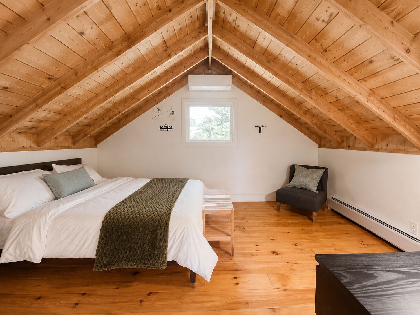 Quarto com teto de madeira inclinado, cama com roupa de cama branca e manta verde, um banquinho, uma cadeira e uma janela pequena.
