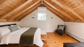 Quarto com teto de madeira inclinado, cama com roupa de cama branca e manta verde, um banquinho, uma cadeira e uma janela pequena.