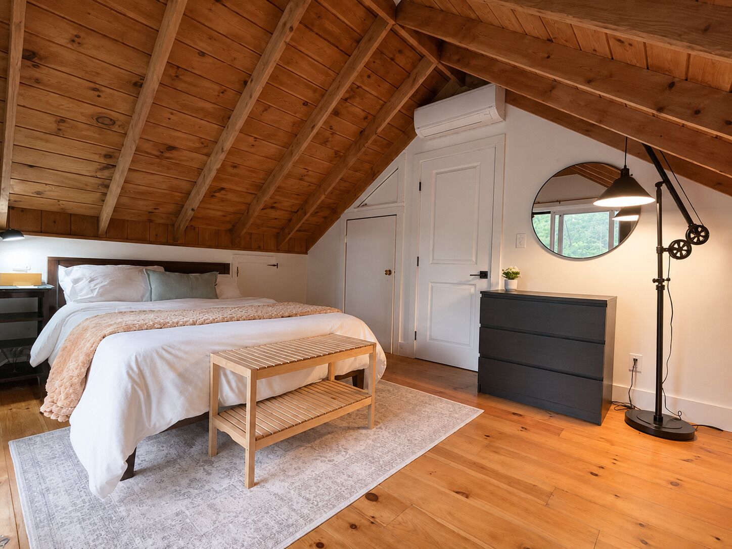 A bedroom with a sloped ceiling, a bed with a bench, a dresser, a mirror, and a floor lamp.