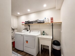 Uma lavanderia com duas máquinas de lavar de carregamento frontal, uma pia e uma prateleira com toalhas dobradas e produtos de limpeza.