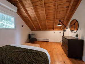 A bedroom with a wooden ceiling, a bed, a dresser, a desk, and a window with a view of trees.