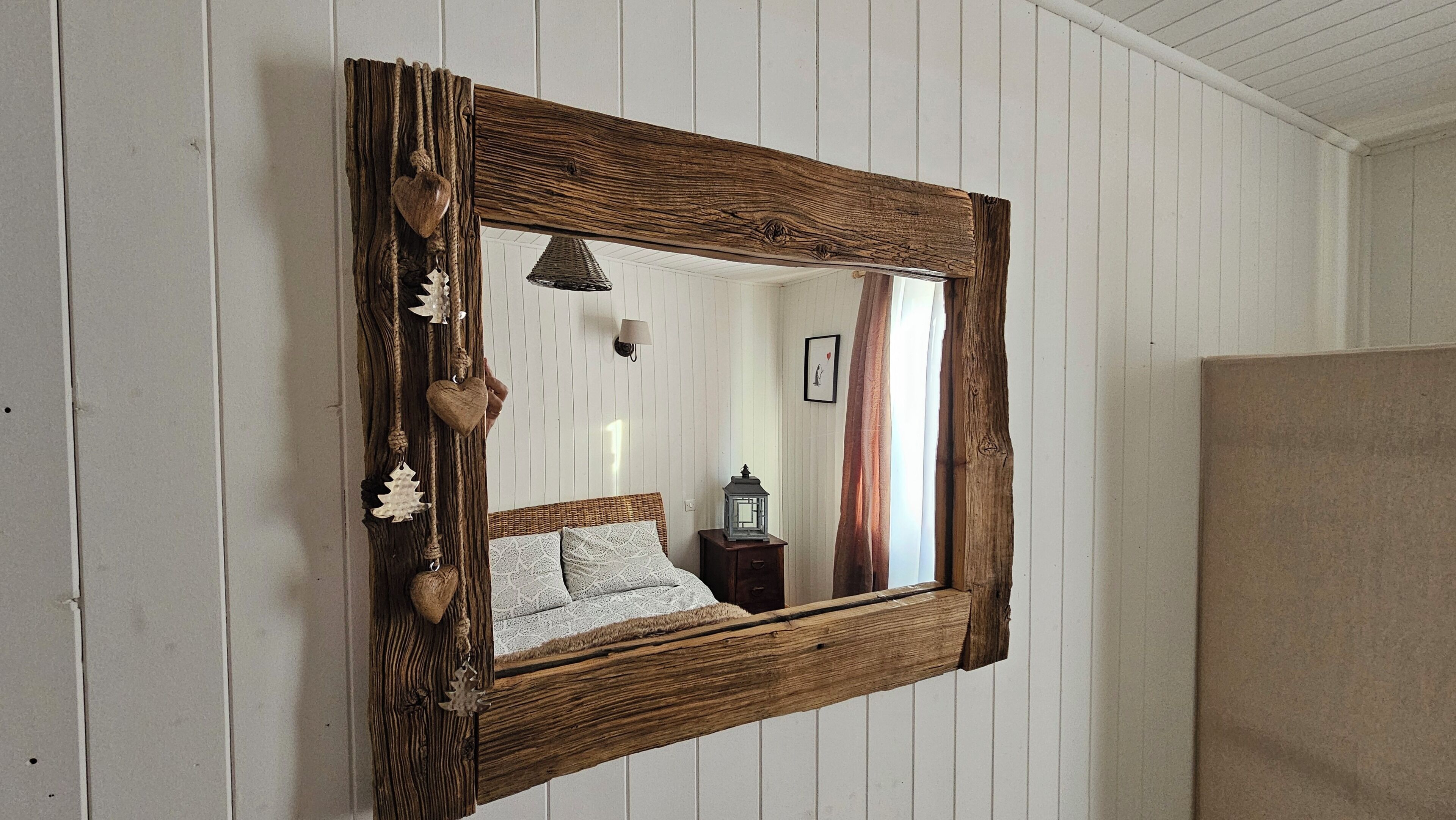 Een rustieke spiegel met houten lijst, waarin een bed en nachtkastje te zien zijn.