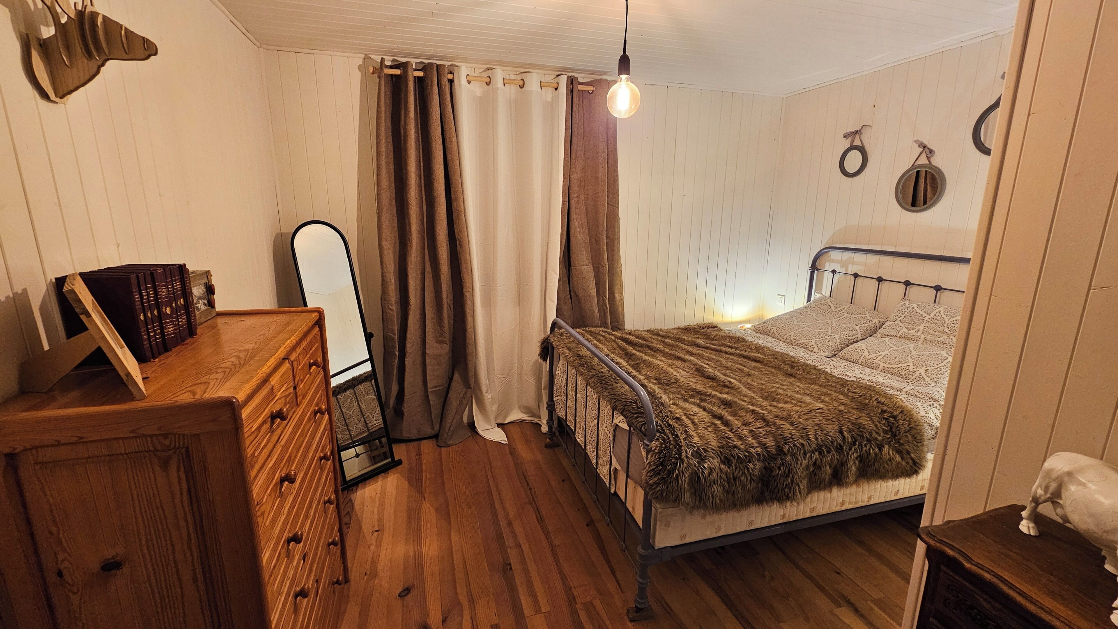 A bedroom with a wooden dresser, a bed with a patterned blanket, a hanging light bulb, and a mirror.