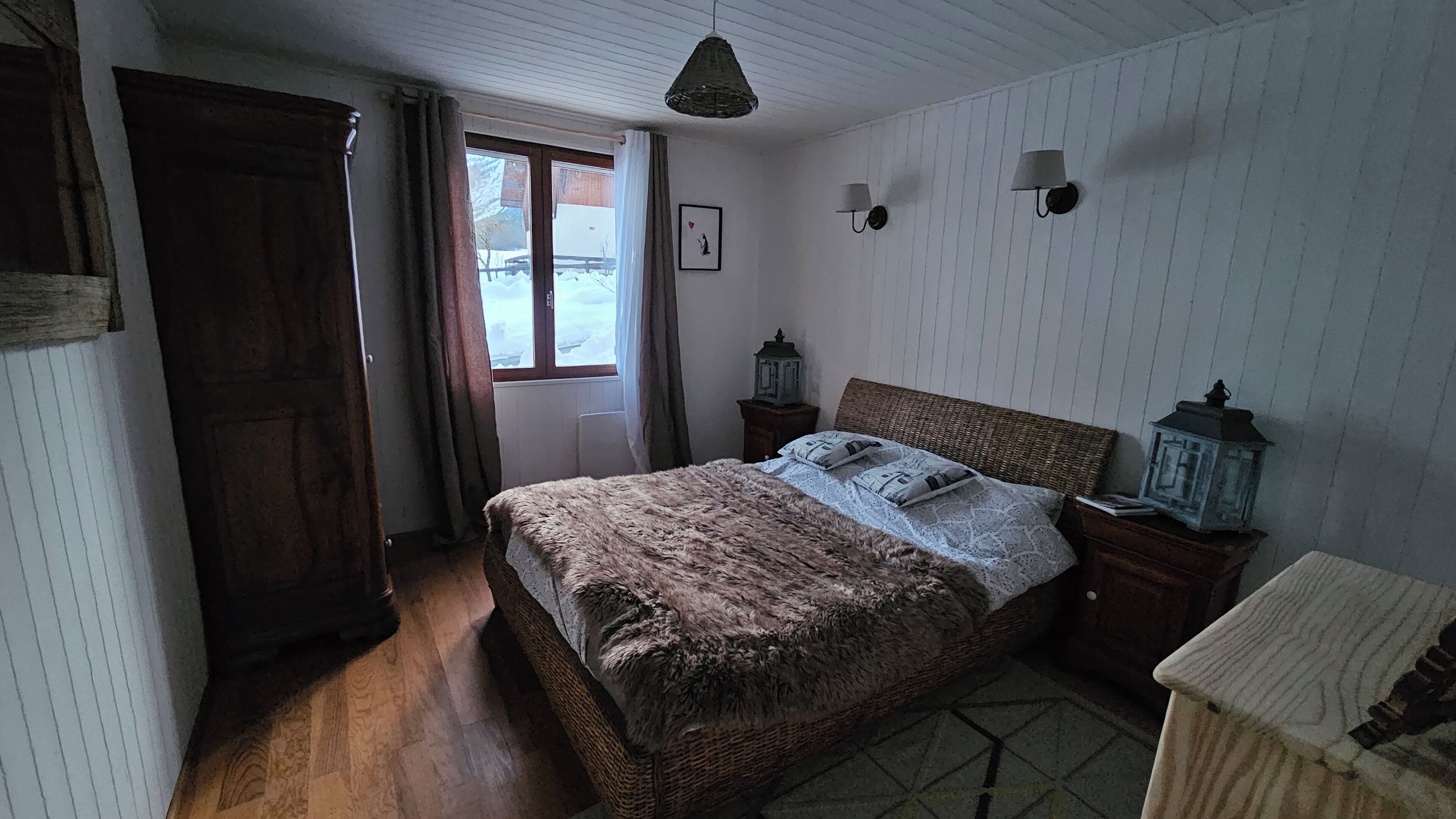 Un dormitorio con cama de madera, ventana con cortinas, lámpara de pared y una pequeña mesa con un farol.