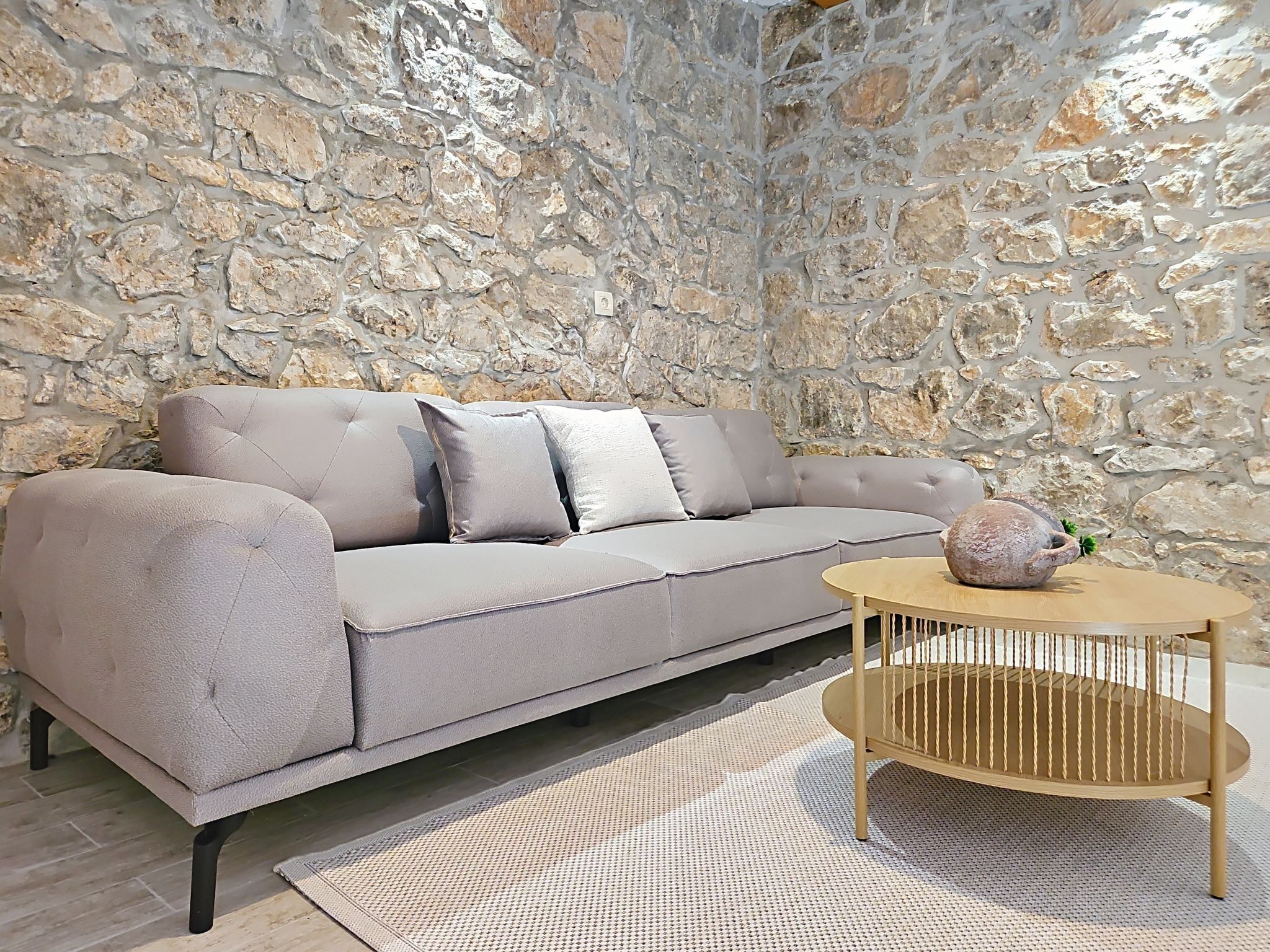 Un canapé beige avec des coussins, adossé à un mur en pierre, une table basse ronde en bois avec un panier en osier, et un tapis à motifs sur un plancher en bois.