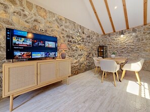 A stone wall interior with a flat-screen TV displaying various streaming services, a wooden cabinet with a woven front, a table with chairs, and a lamp. - Vacation home Ana by Interhome (Trilj)