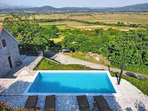 A swimming pool with lounge chairs, a stone building, and a view of a rural landscape with fields and houses. - Vacation home Marija by Interhome (Trilj)