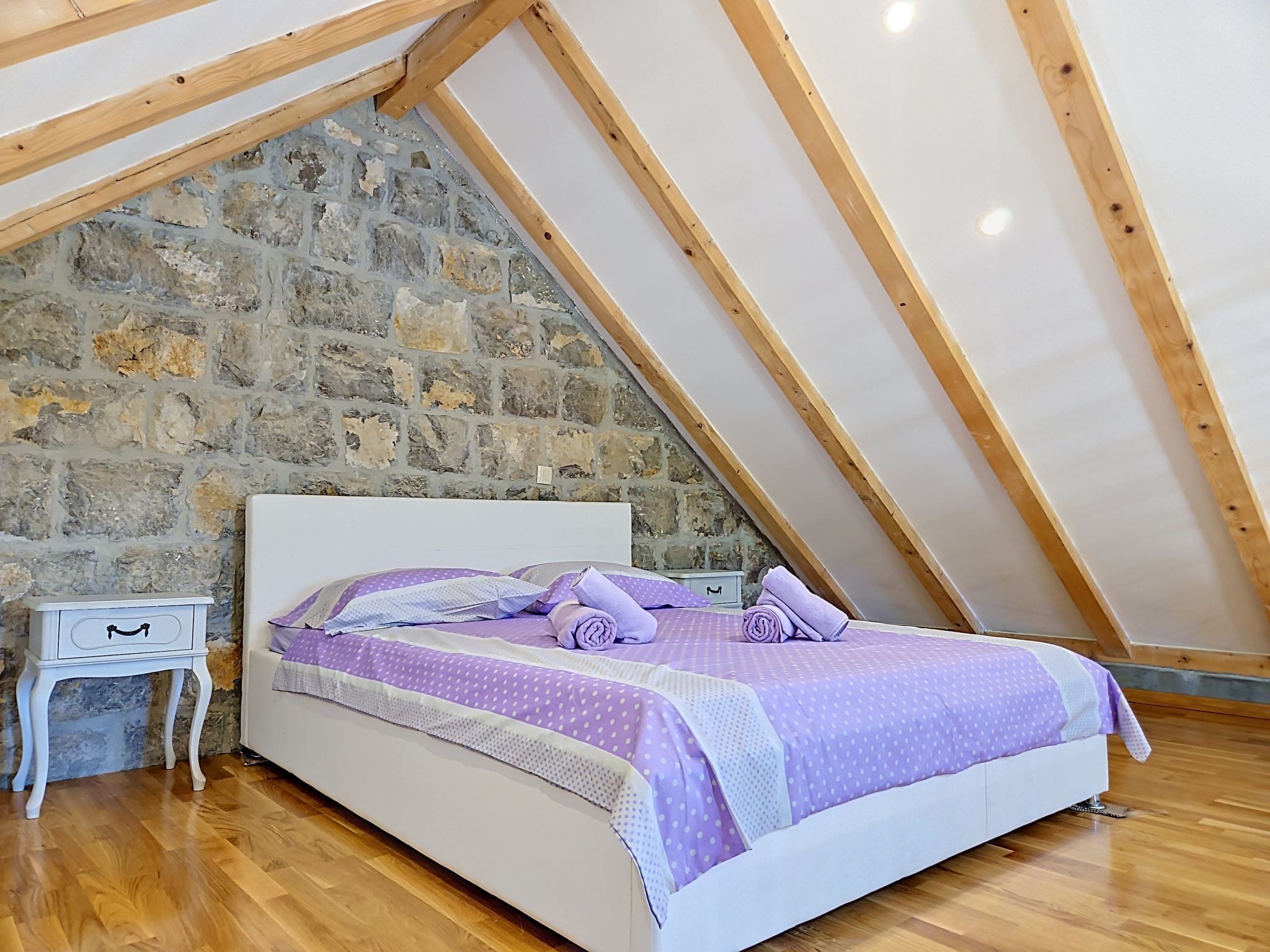 A bedroom with a stone wall, a wooden ceiling, a bed with purple bedding, and a nightstand.