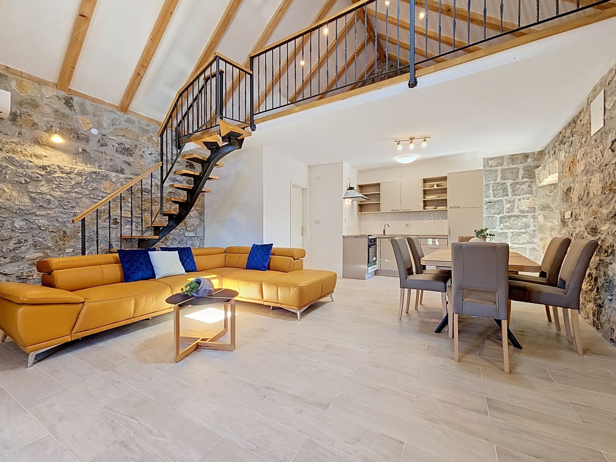 A modern living room with a curved staircase, a sectional sofa, a dining area with a table and chairs, and a kitchen in the background.
