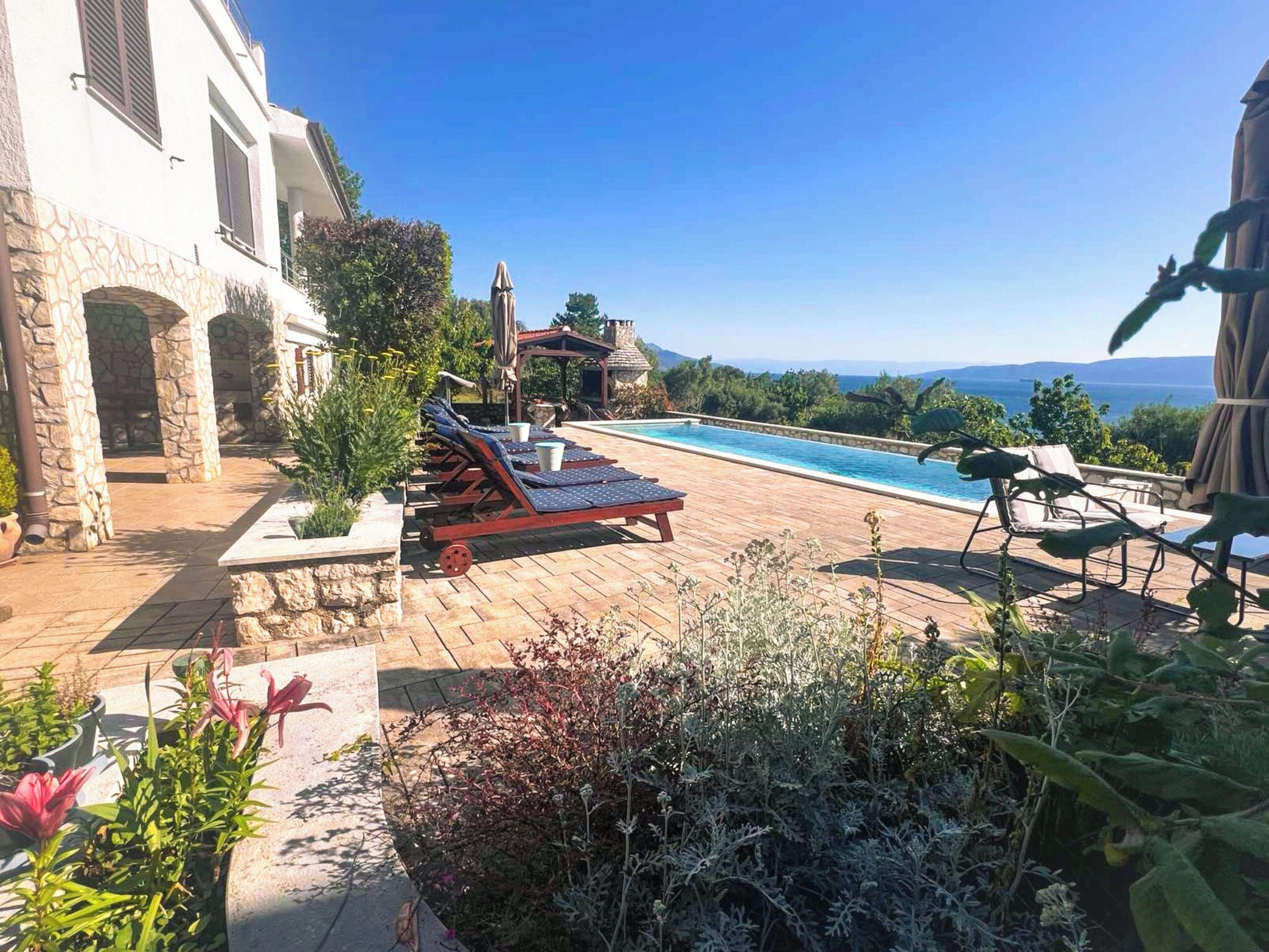 Un espace aménagé au bord de la piscine, avec des chaises longues, un mur en pierre et une vue sur la mer.