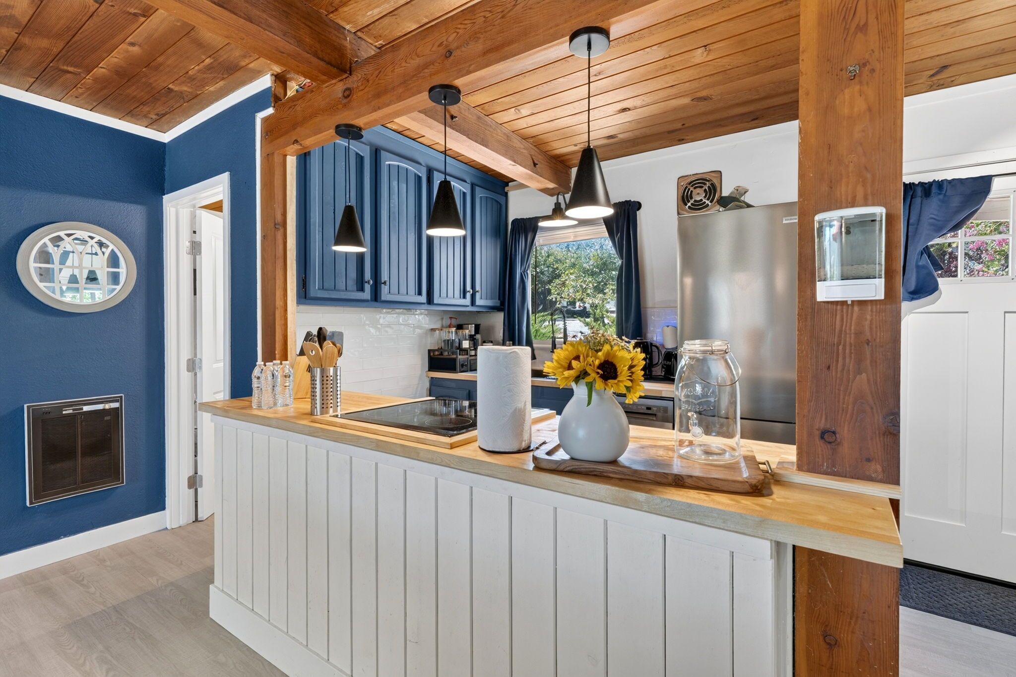 Une cuisine avec des armoires bleues, des poutres en bois au plafond, des luminaires suspendus, un îlot central orné d’un vase de tournesols et un réfrigérateur en acier inoxydable.