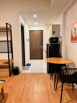 A compact room with a wooden floor, a small dining table with chairs, a kitchenette, a door leading to another room, and a wall-mounted artwork. - Soho residence studio apartment (Ho Chi Minh City)