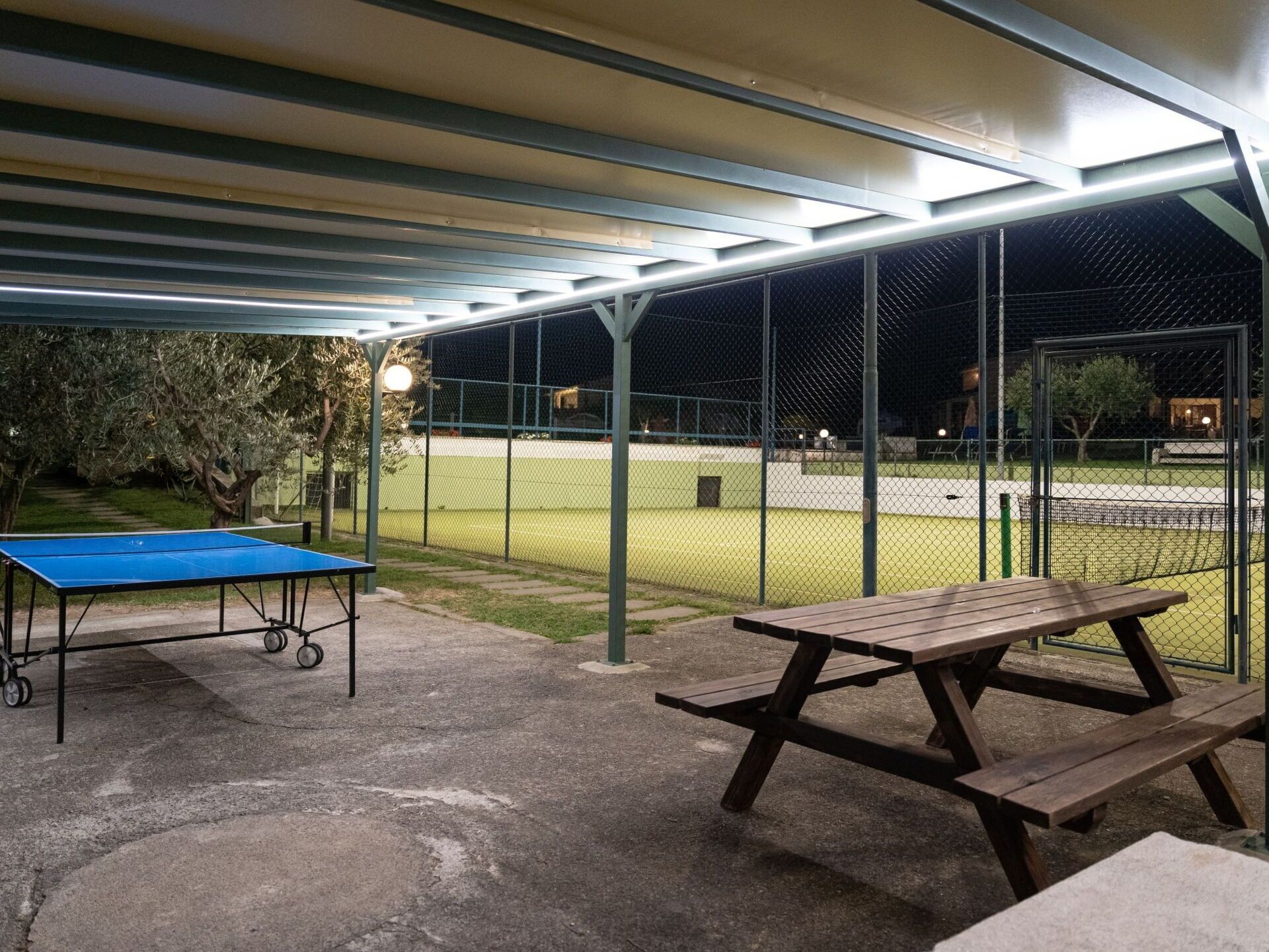 An outdoor tennis court with a covered seating area, a ping pong table, and a picnic table.