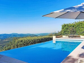 En infinitypool med fri utsikt över avlägsna berg, en uteplats med ett bord och stolar samt ett stort parasoll som ger skugga.