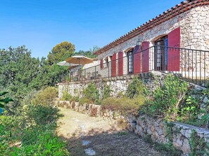 Un bâtiment en pierre avec des volets rouges, un balcon doté d’une rambarde en métal et un jardin agrémenté de diverses plantes.