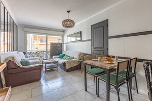 A modern living room with a sofa, a dining table with chairs, a hanging light fixture, and a view through a window. - Design 2BR | Private Parking, Beach & Train (Golfe Juan Vallauris)
