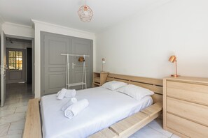 A bedroom with a bed, a wooden dresser, a nightstand, a hanging light fixture, and a door with a towel rack. - Design 2BR | Private Parking, Beach & Train (Golfe Juan Vallauris)