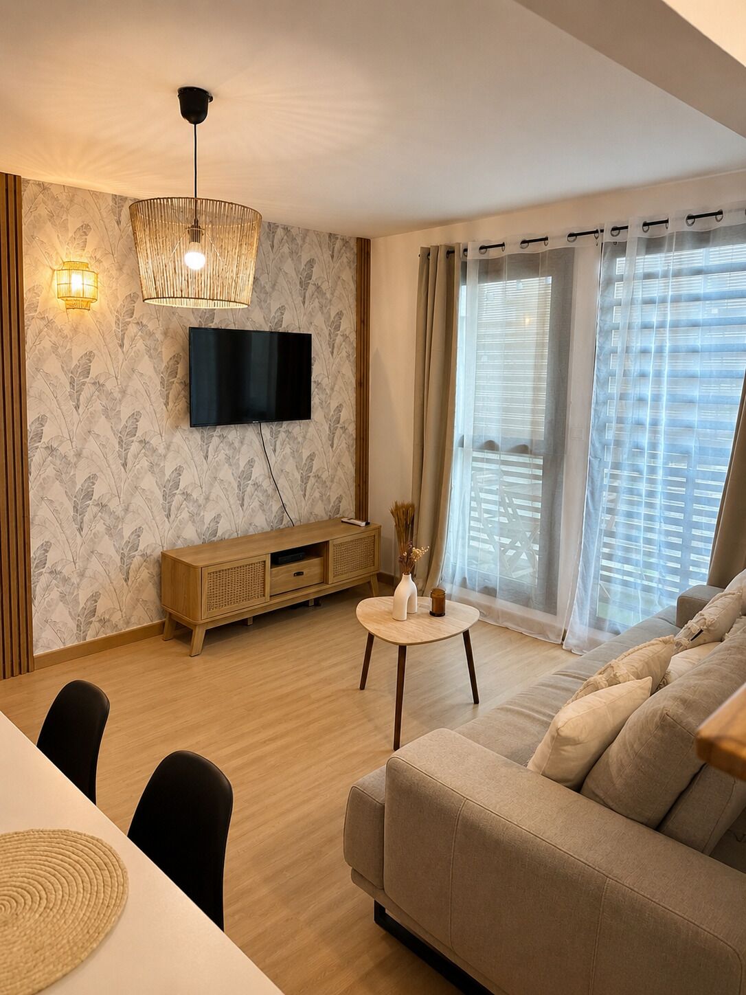A modern living room with a flat-screen TV, a wooden TV stand, a round coffee table, a beige sofa, and a dining area with a white table and black chairs.