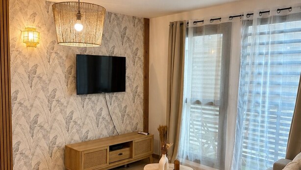 Una sala moderna con una televisión de pantalla plana, un mueble de televisión de madera, una mesa de centro redonda, un sofá color beige y un comedor con una mesa blanca y sillas negras.
