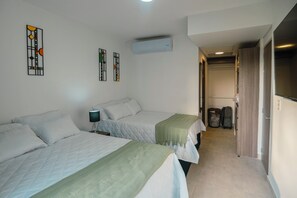Ein Hotelzimmer mit zwei Betten, einem Nachttisch mit Lampe, einer Wand-Klimaanlage und einem Flur, der zu einem Badezimmer führt.