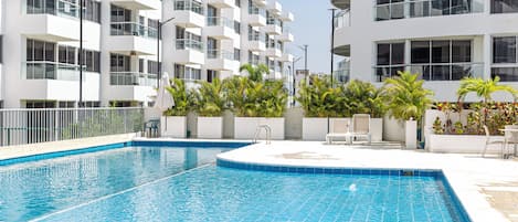 A swimming pool with a tiled edge, surrounded by a white building with balconies and a clear sky.