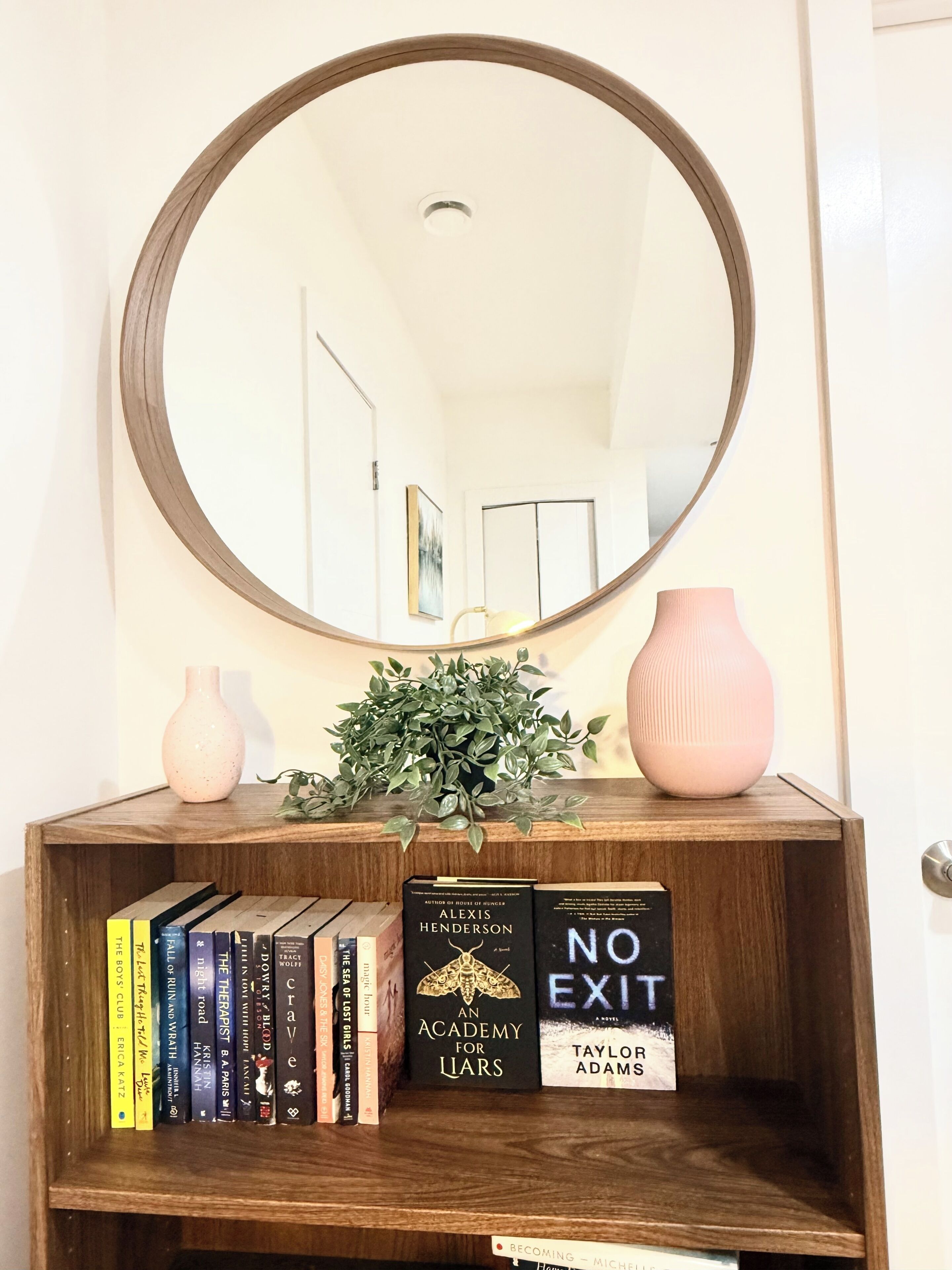 A wooden console table with a mirror above it, a potted plant, a pink vase, and a bookshelf with various books.