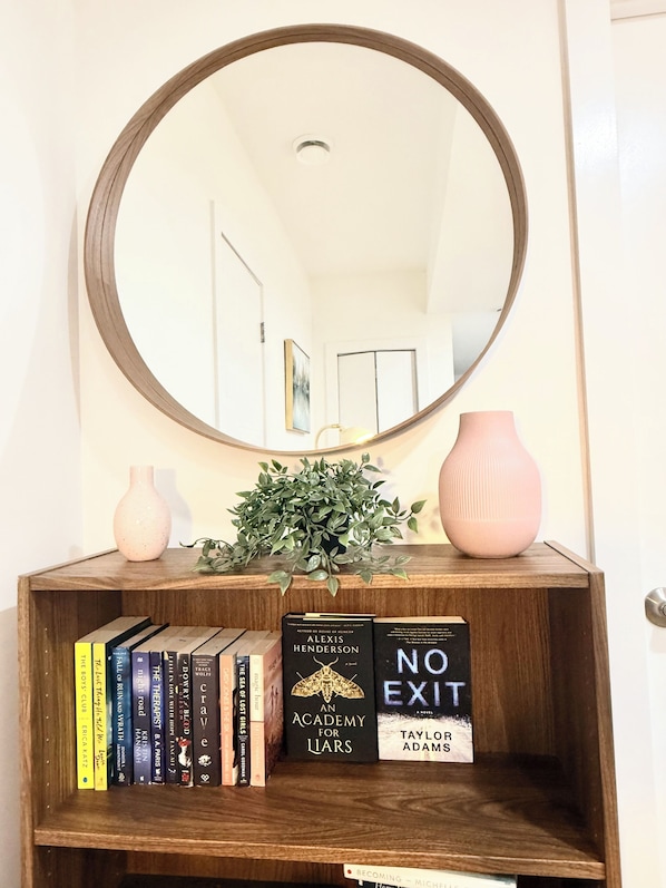 A wooden console table with a mirror above it, a potted plant, a pink vase, and a bookshelf with various books. - Modern 2BR suite | U of A •Whyte Ave •Downtown (Edmonton)