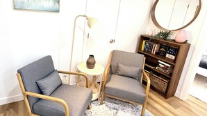 Een kamer met twee grijze fauteuils, een klein rond tafeltje met een vaas, een vloerlamp, een boekenkast vol boeken en een schilderij aan de muur.