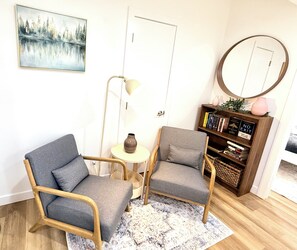 A room with two grey armchairs, a small round table with a vase, a floor lamp, a bookshelf with books, and a painting on the wall. - Modern 2BR suite | U of A •Whyte Ave •Downtown (Edmonton)