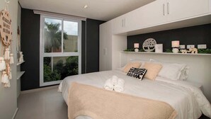Un dormitorio con un ventanal grande, una cama con ropa de cama blanca, una estantería con objetos decorativos y un letrero sujeto a la pared.