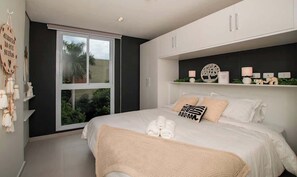 Ein Schlafzimmer mit großem Fenster, einem Bett mit weißen Leinen, einem Regal mit Dekoration und einem an der Wand befestigten Schild.