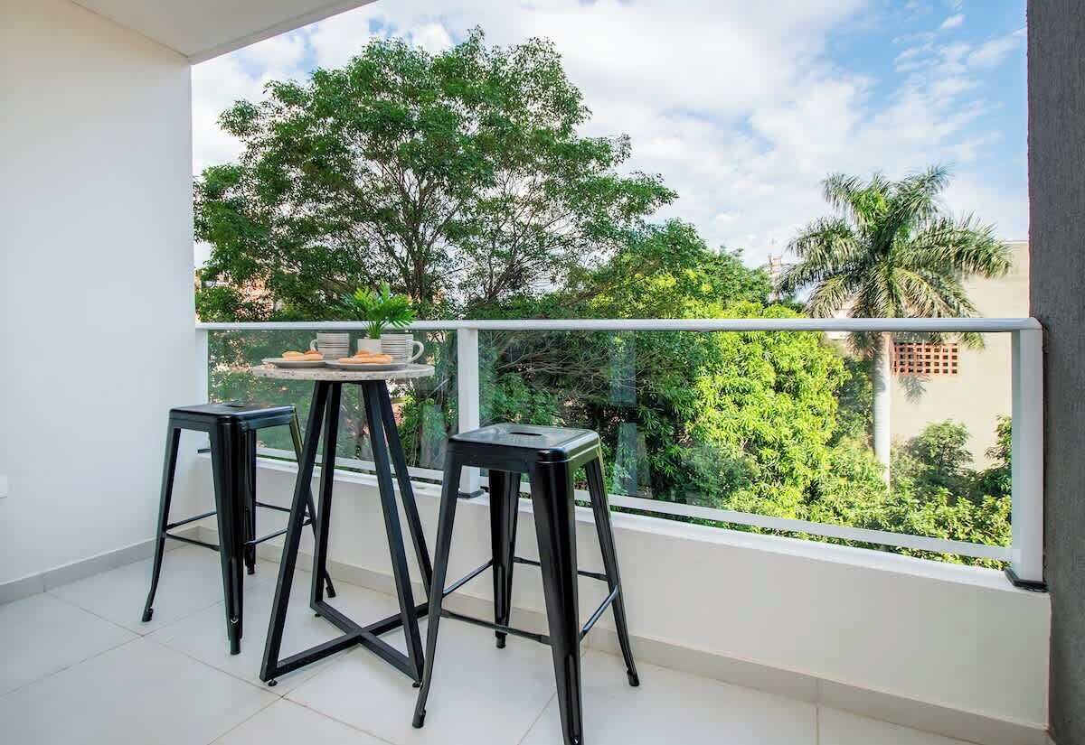 Un balcón con dos taburetes negros, una mesa, barandilla de cristal y vista a árboles y edificios.