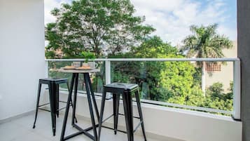 Un balcón con dos taburetes negros, una mesa, barandilla de cristal y vista a árboles y edificios.