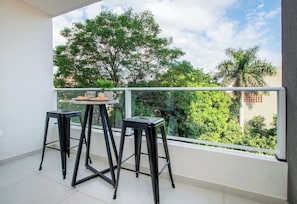 Un balcó amb dos tamborets negres i una taula, una barana de vidre i vista a arbres i edificis.