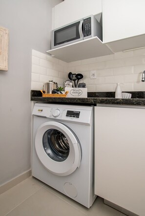 A compact laundry area with a front-loading washing machine, a microwave above it, and a small countertop with kitchen utensils. - Corporate Stays First Living - 106 (Asunción)