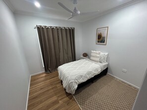 Une chambre à coucher comprenant un lit, une table de chevet, un ventilateur de plafond, une fenêtre avec des rideaux et un tableau encadré accroché au mur.