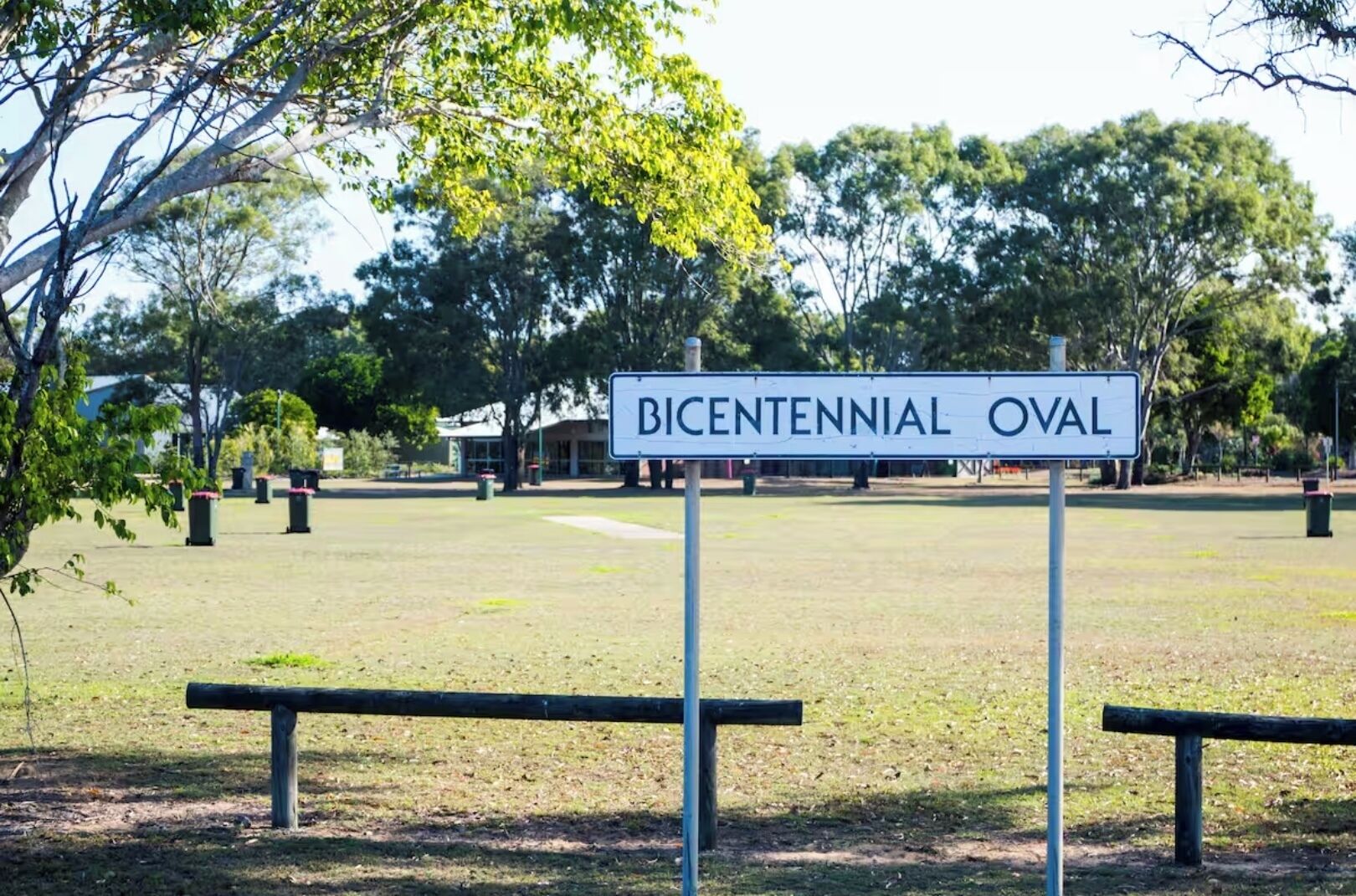 Placa com a inscrição 'BICENTENNIAL OVAL' em um parque com gramado, árvores e bancos.