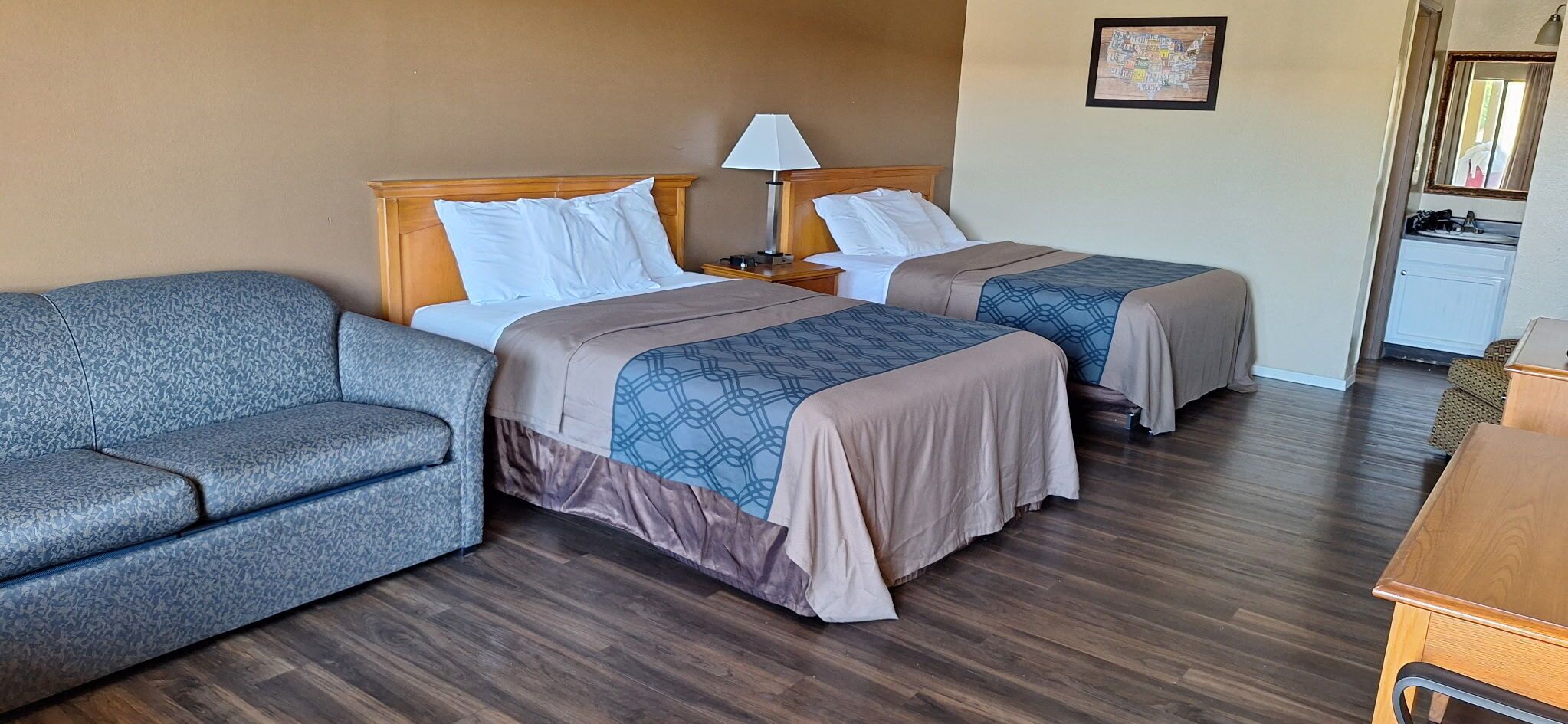 A hotel room with two beds, a sofa, a nightstand with a lamp, and a small desk with a mirror.