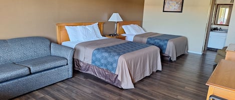 A hotel room with two beds, a sofa, a nightstand with a lamp, and a small desk with a mirror.