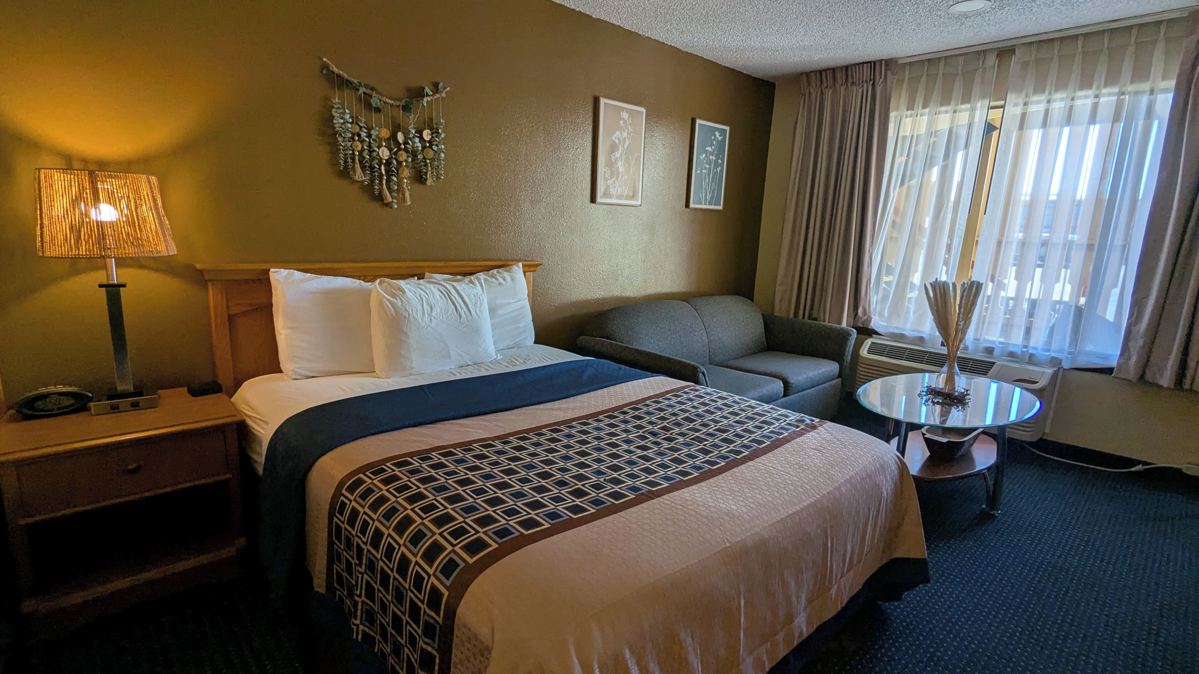 A hotel room with a bed, a desk, a chair, a small table, and a window with curtains.