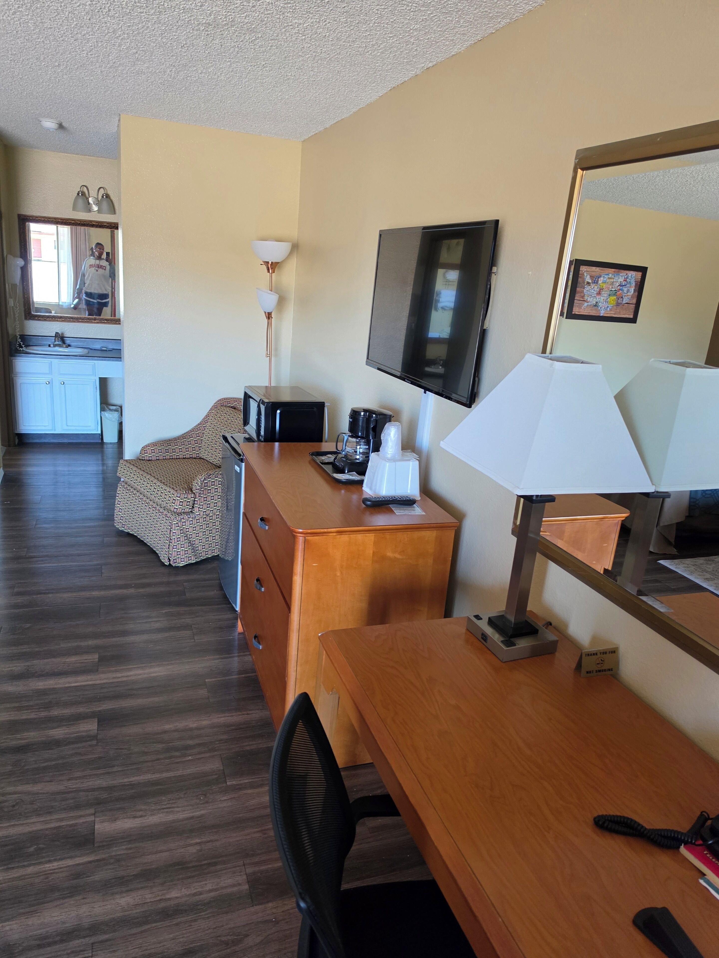 A hotel room with a wooden desk, a television, a microwave, a coffee maker, a chair, a dresser, and a small table with a lamp.
