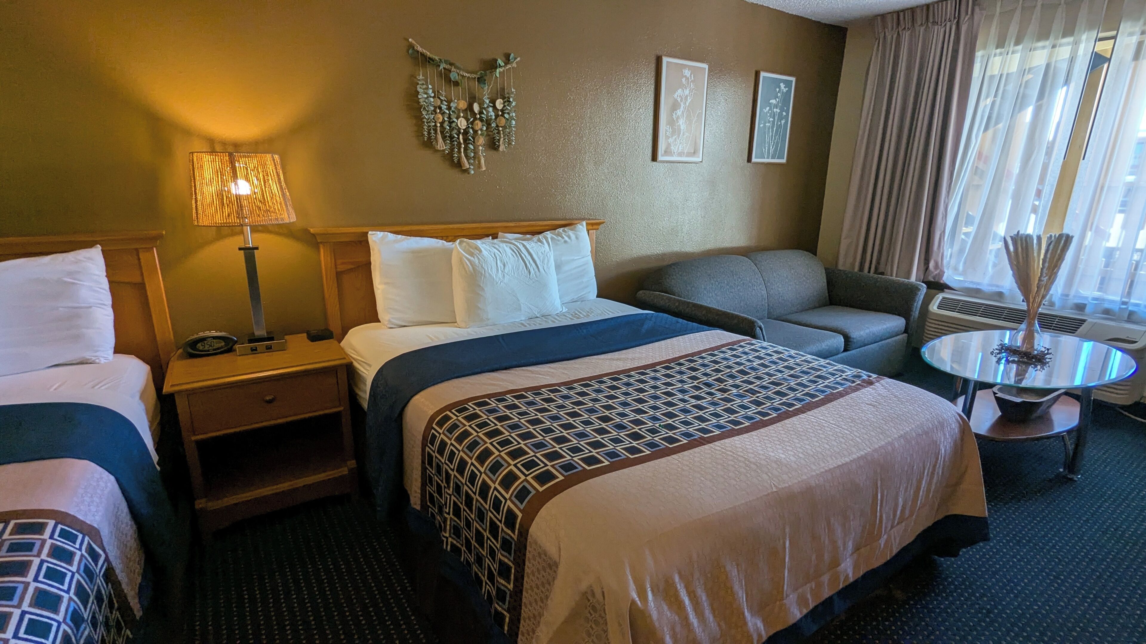A hotel room with two beds, a sofa, a small table with a vase, and a wall-mounted decoration.