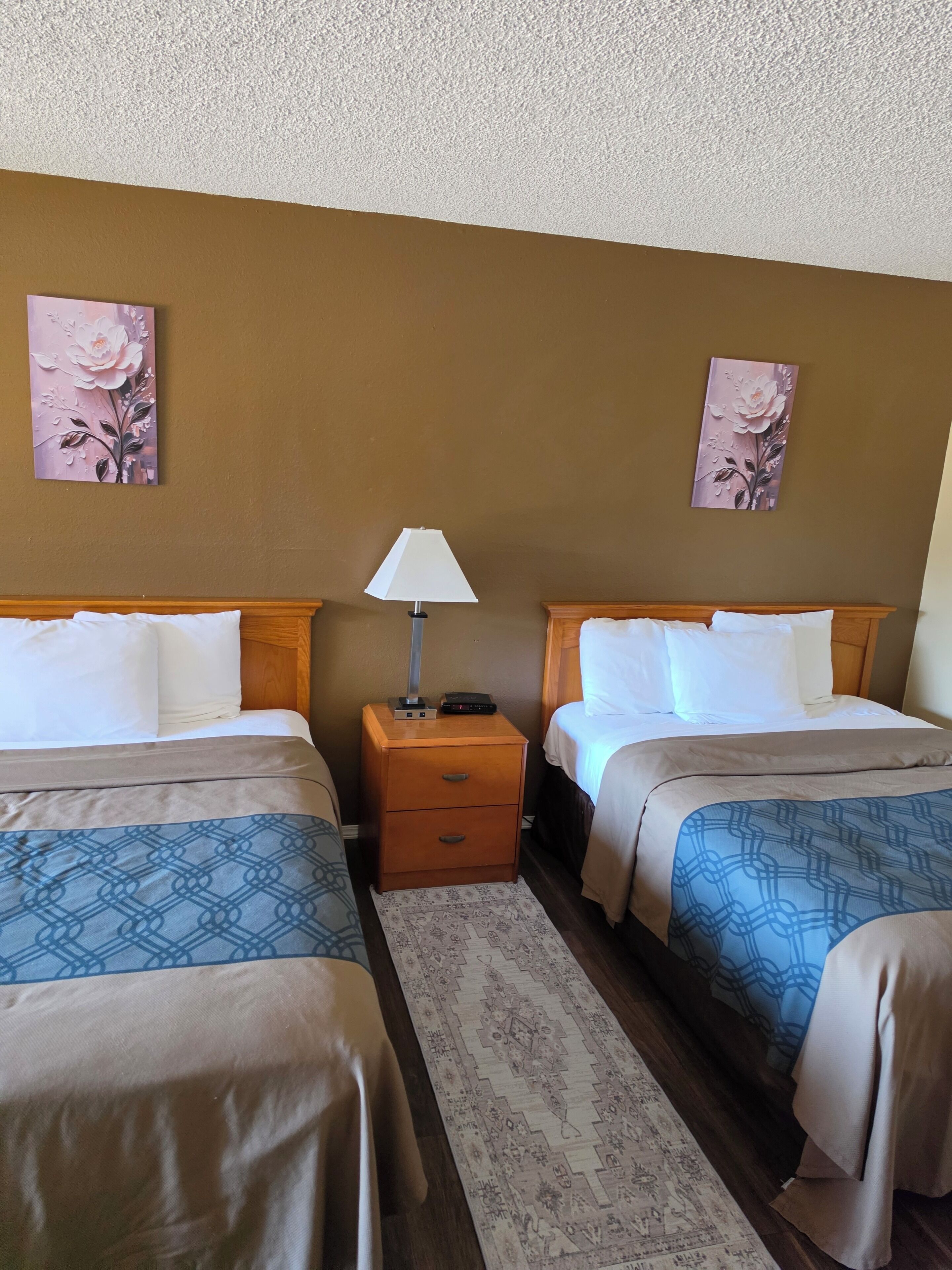 A hotel room with two beds, a nightstand with a lamp, and two floral wall art pieces.