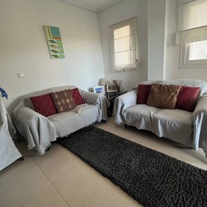 A living room with two sofas, a rug, a table with a lamp, and a window with blinds. - Adriatico Groundfloor Frontline Golf - 2 Bath - 3 Min Walk to Amenities (Sucina)