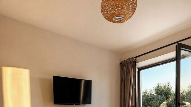 A bedroom with a TV, a bed with a blanket, a bedside table with a vase, and a view of the outdoors. - Conte Maris II - Sea View Mediterranean Escape (Tragaki)