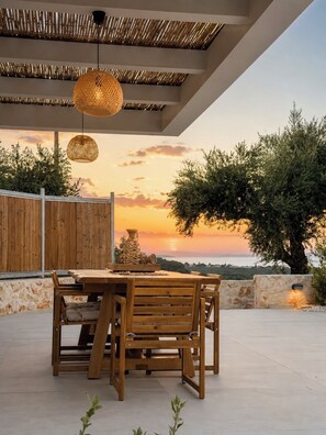 An outdoor dining area with a wooden table and chairs, a woven ceiling, and a view of the sea at sunset. - Conte Maris II - Sea View Mediterranean Escape (Tragaki)