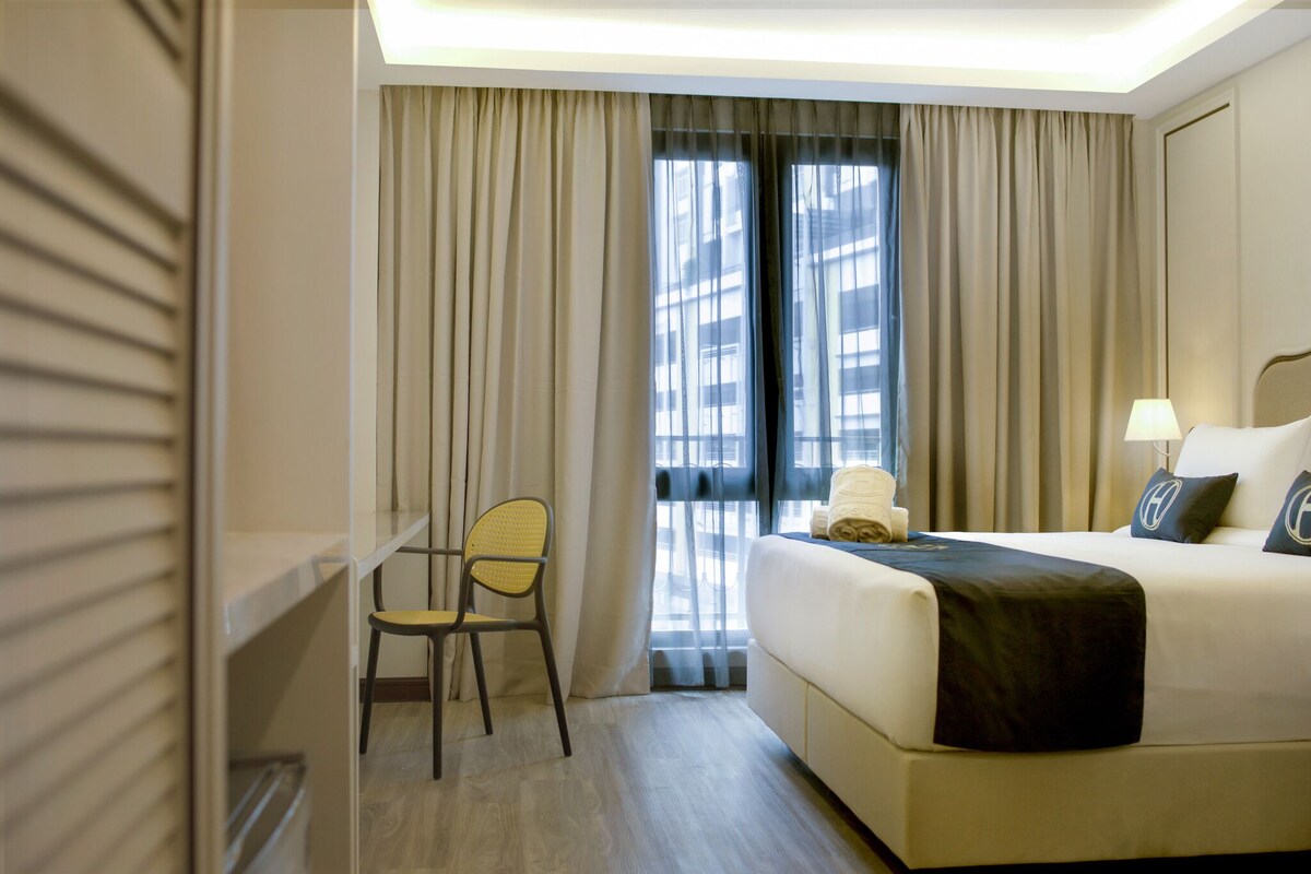 Una habitación de hotel con cama, una silla, un escritorio y un ventanal con cortinas.