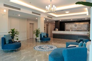 Un vestíbulo de hotel con una recepción, sofás azules, una araña de cristal y suelo de mármol.