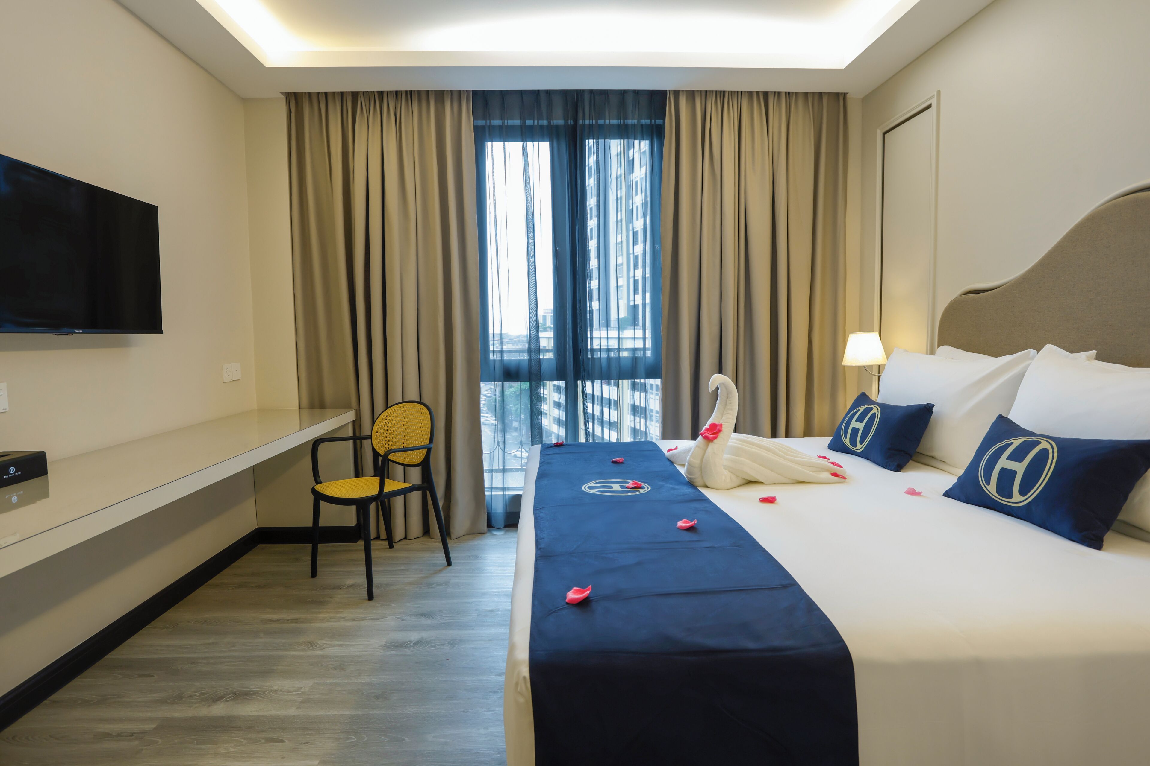 A hotel room with a bed, a desk, a chair, a TV, and a view of buildings through the window.