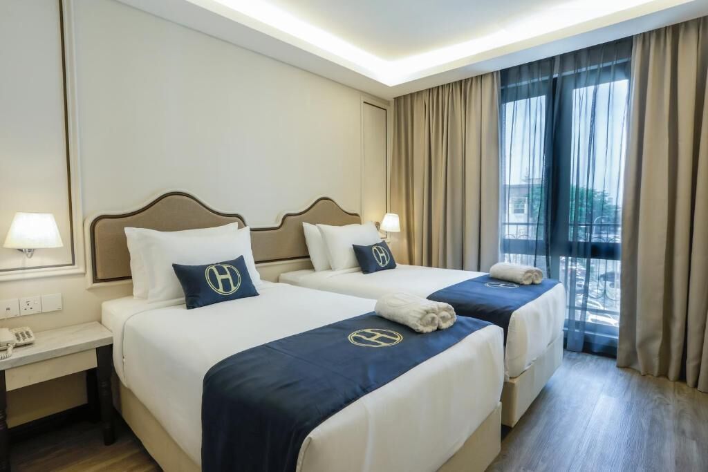 A hotel room with two beds, each with a headboard, bedside tables with lamps, and a view of the outdoors through large windows.